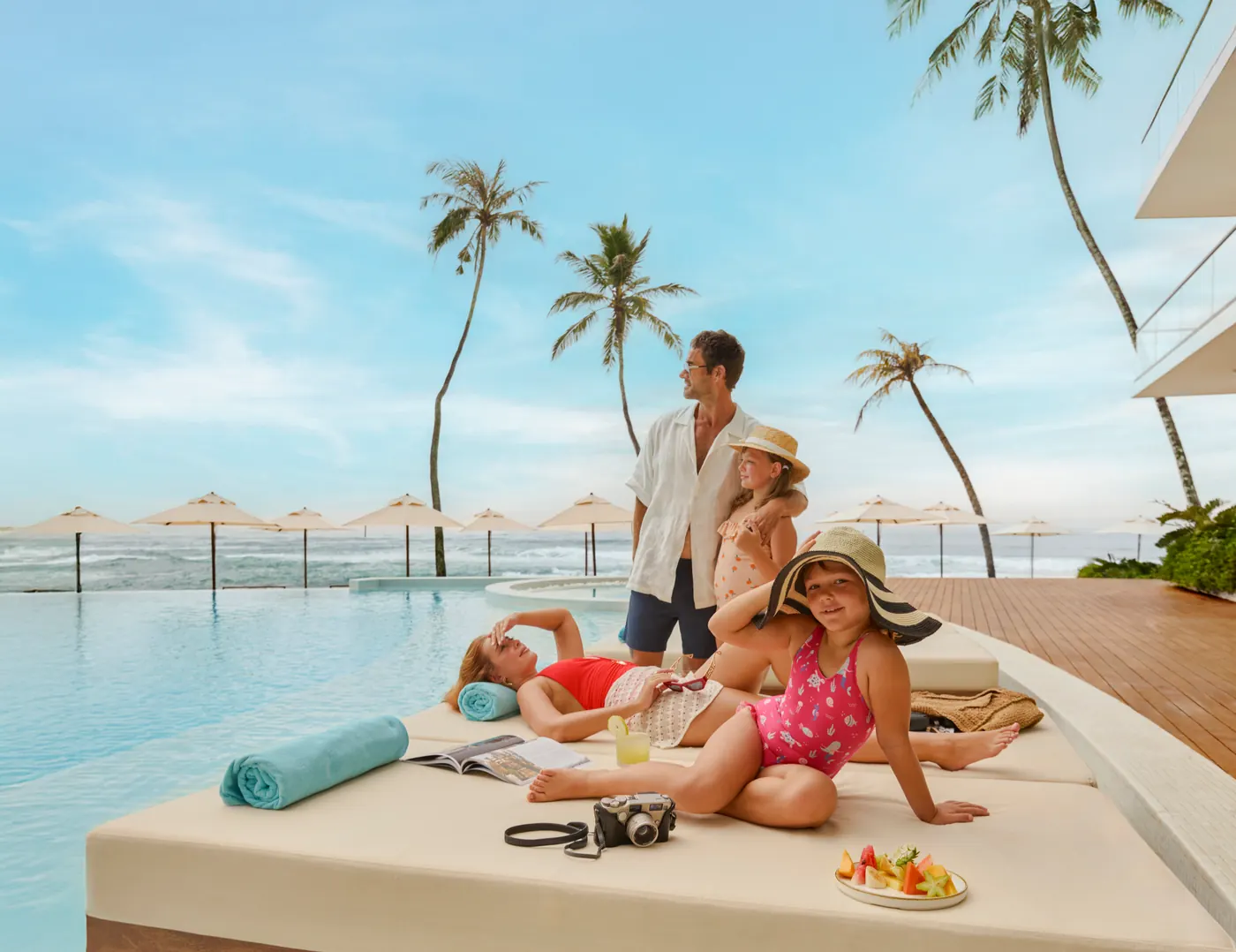 A family on a poolside daybed with a fruit plate and a camera, palm trees and the ocean behind them